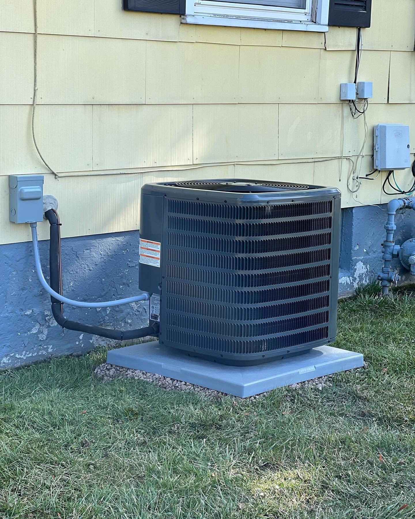 A residential outdoor air conditioning unit is installed next to a yellow house, sitting on a concrete pad above the grass.
