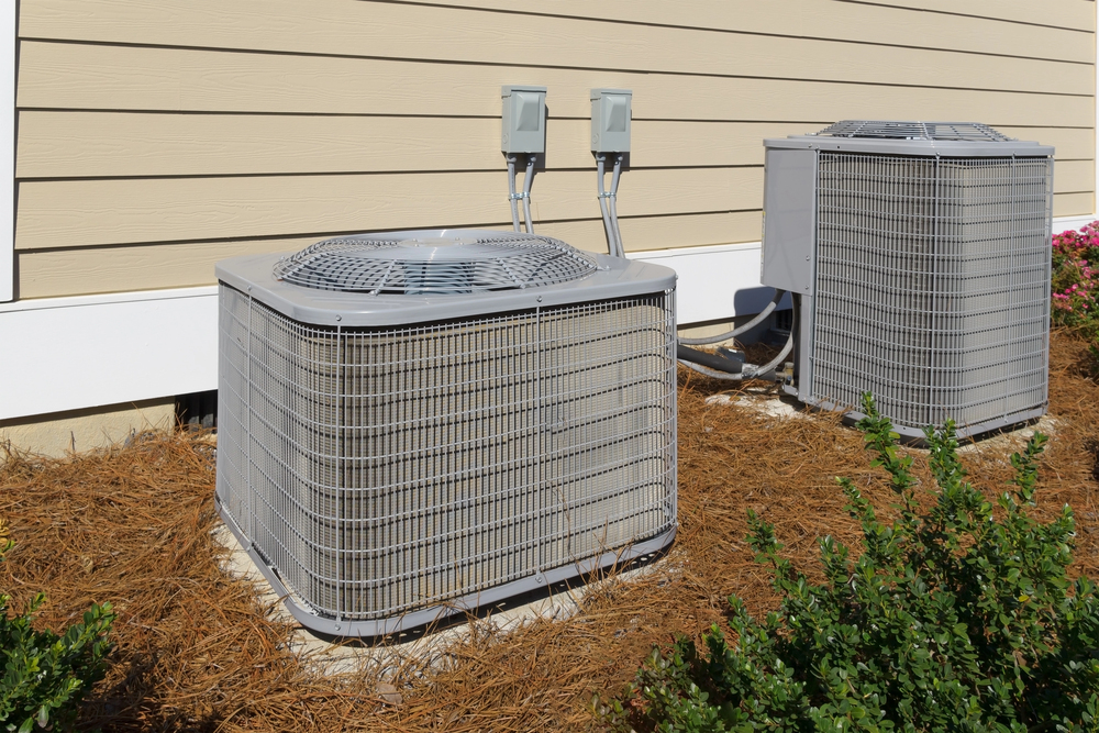 Two outdoor air conditioning units installed beside a beige house, with electrical boxes mounted on the wall and mulch covering the ground.