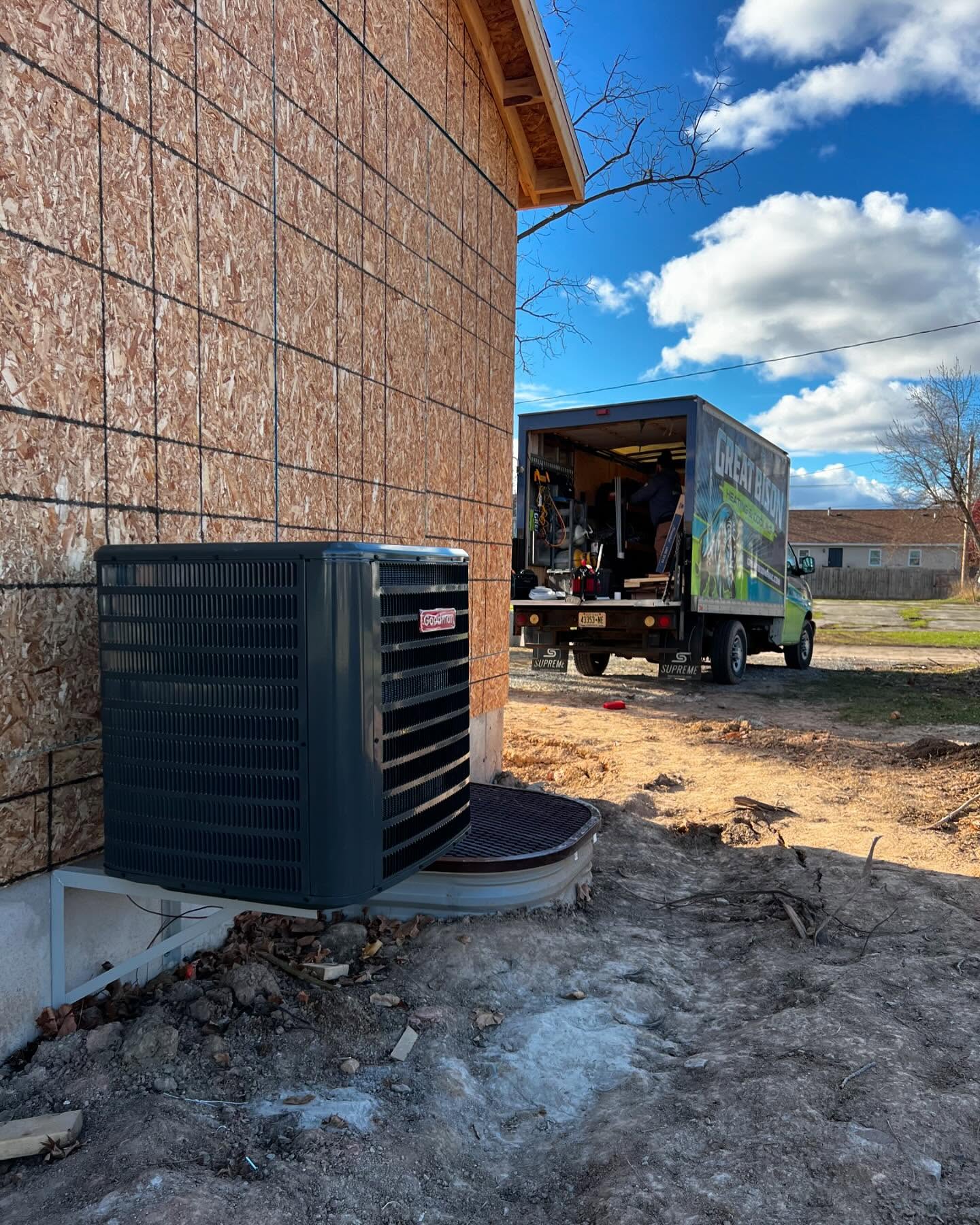 A newly installed HVAC unit is positioned outside a house under construction, with a work truck and tools visible in the background on a clear day.