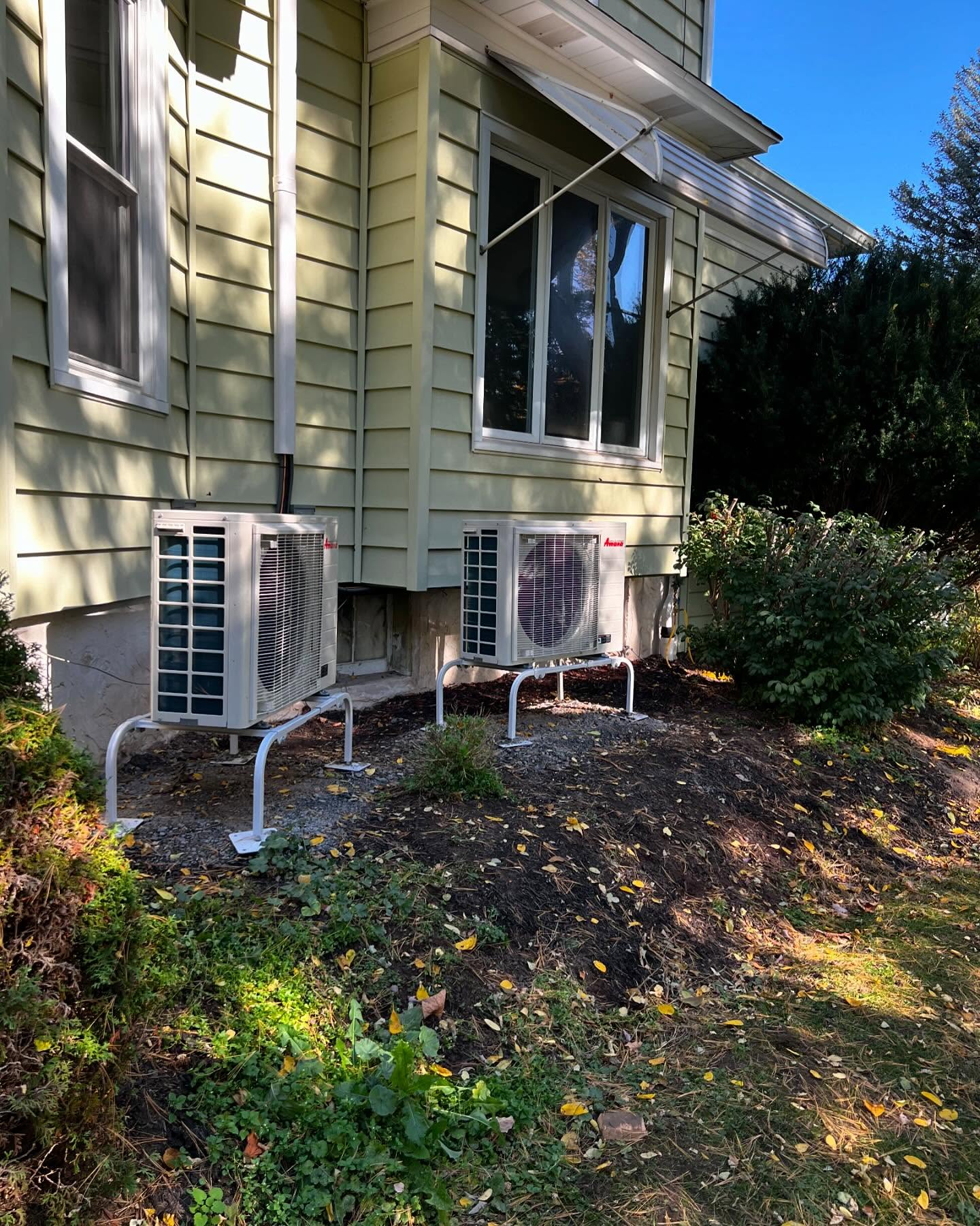 Two outdoor air conditioning units mounted on metal stands are positioned next to the exterior wall of a yellow house, with shrubs and garden beds nearby.