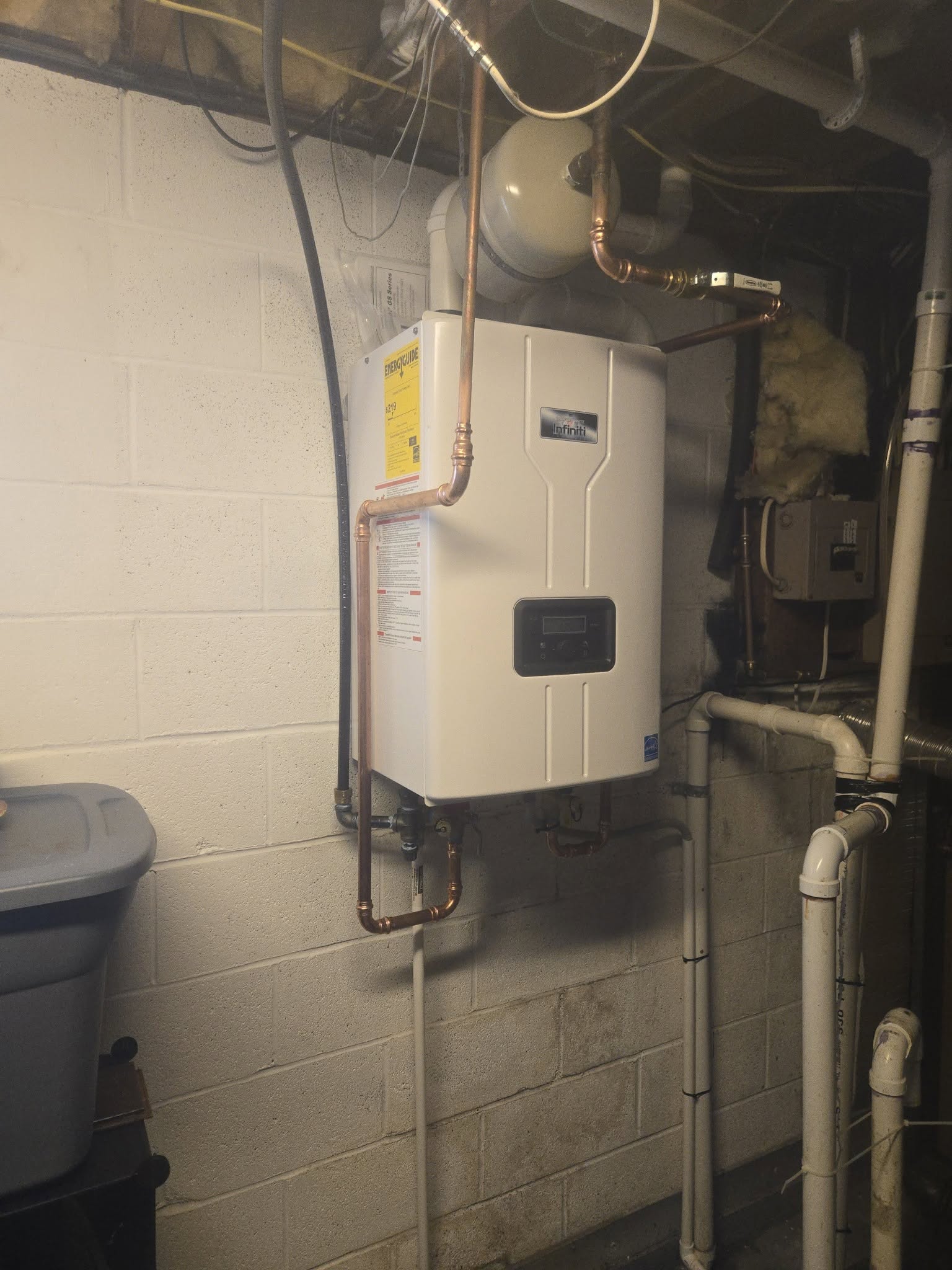 A tankless water heater is mounted on a basement wall, with copper pipes and various plumbing connections visible around it.