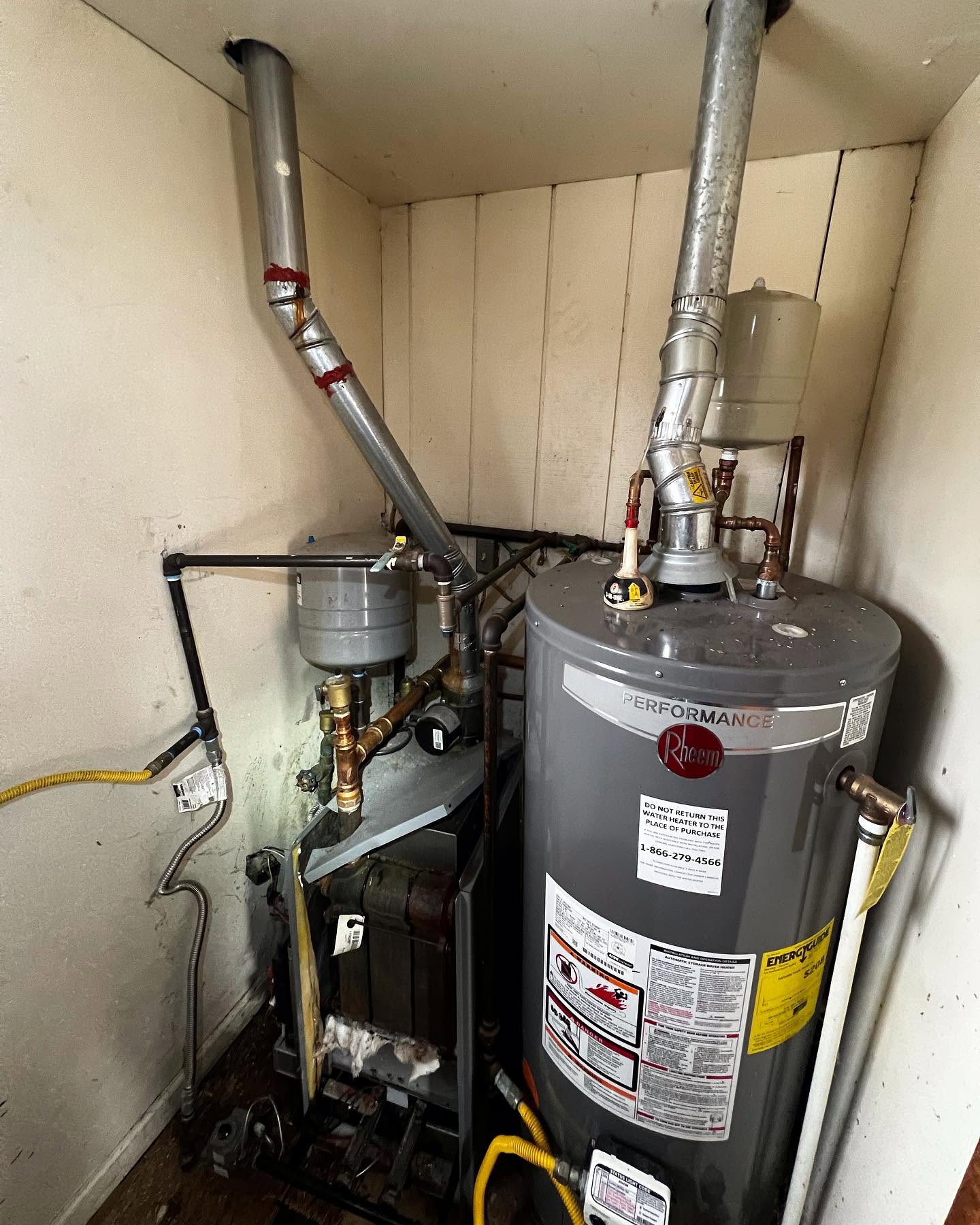 Two water heaters with various pipes and gauges are installed in a small, enclosed utility space with white paneled walls.
