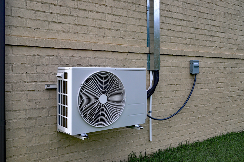 A white outdoor air conditioning unit is mounted on a beige brick wall with electrical wiring running into a nearby gray utility box above grass.
