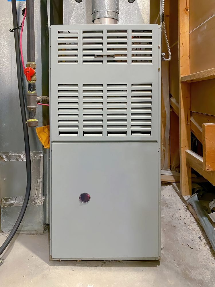 A gray metal furnace unit installed in a utility room with pipes and wires visible on the sides.