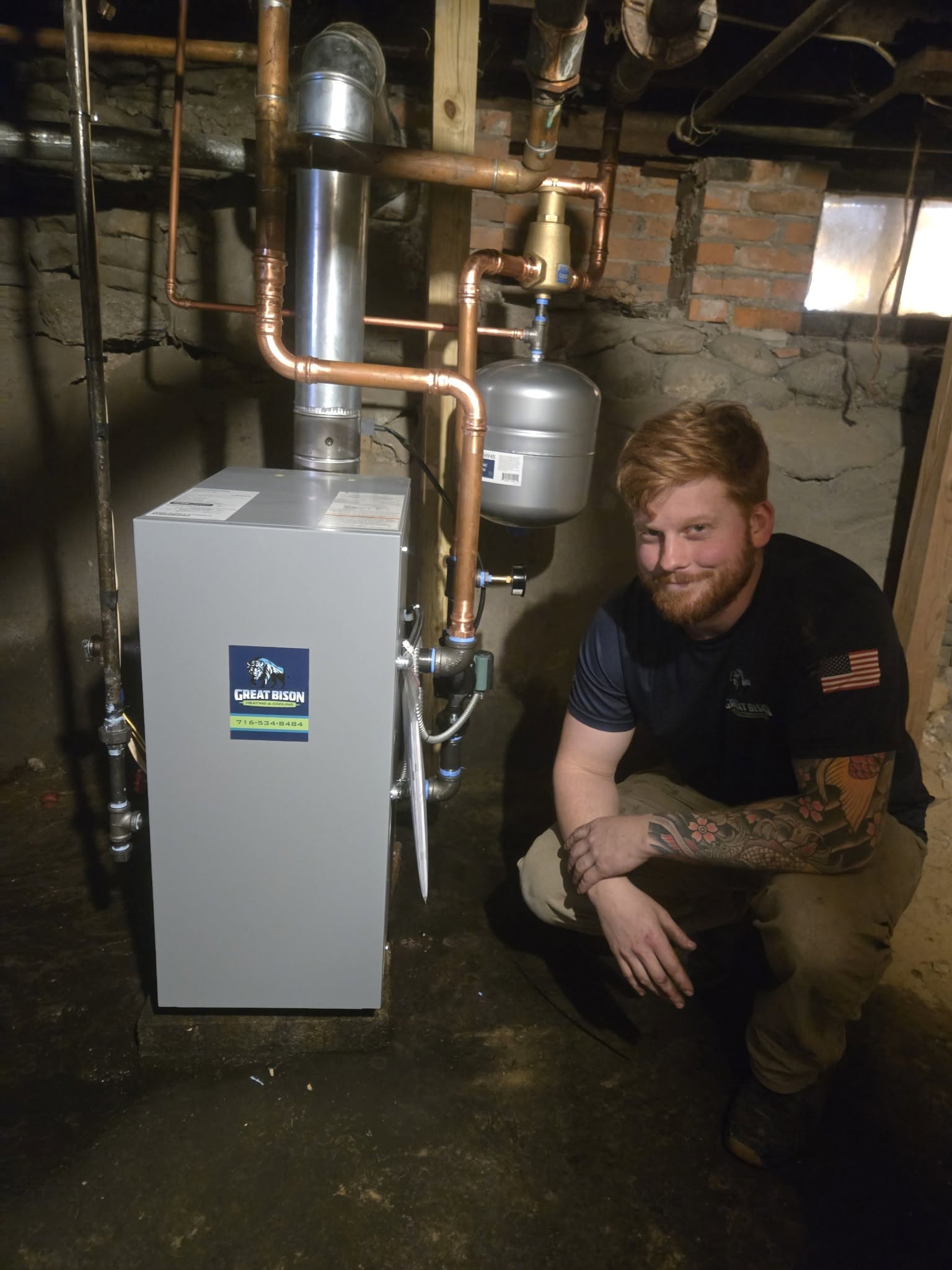 A man kneels next to a newly installed furnace with copper piping in a dimly lit basement with exposed brick and stone walls.