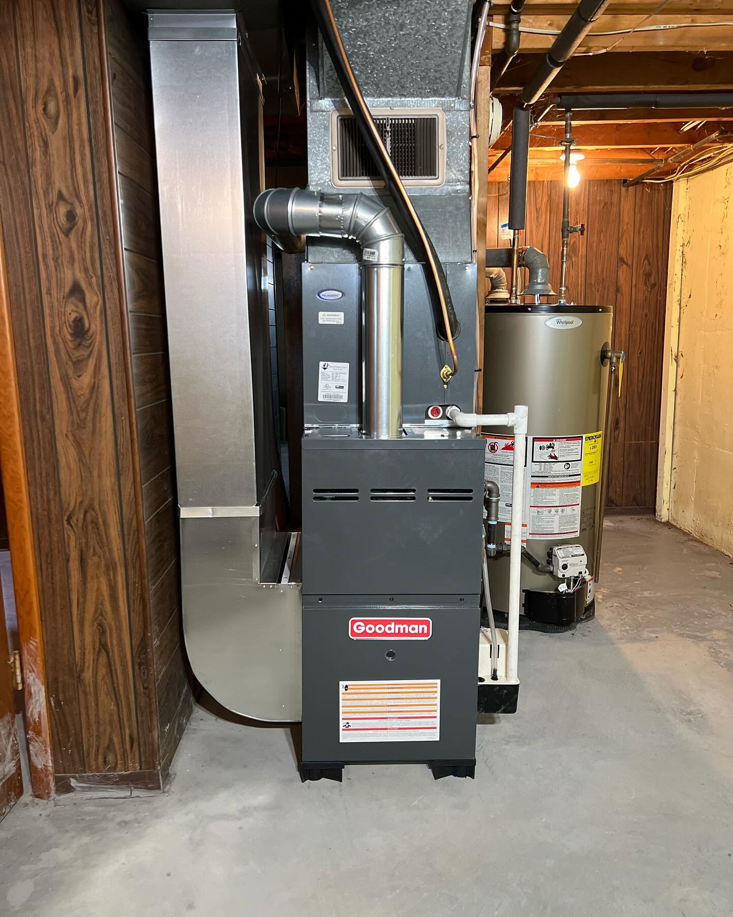 A Goodman furnace and a water heater installed in a basement with wood panel walls and exposed ceiling beams.
