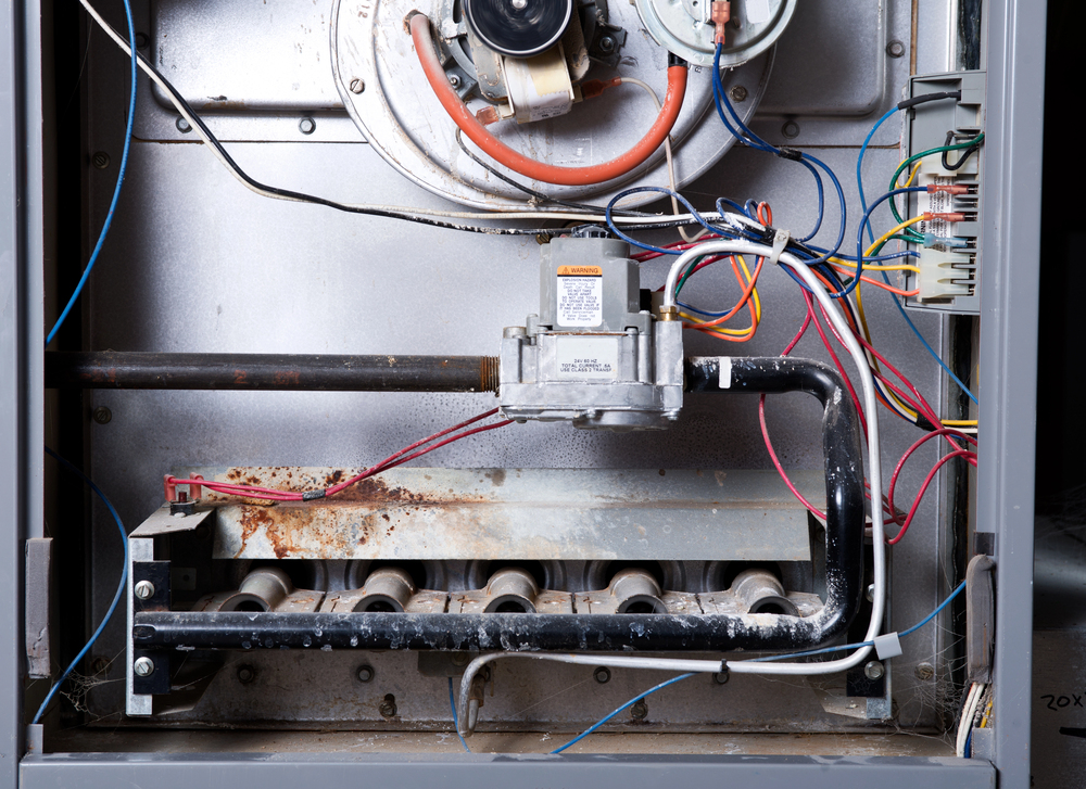 Interior view of a gas furnace showing burners, pipes, wiring, and control components, with some visible rust on the metal surfaces.