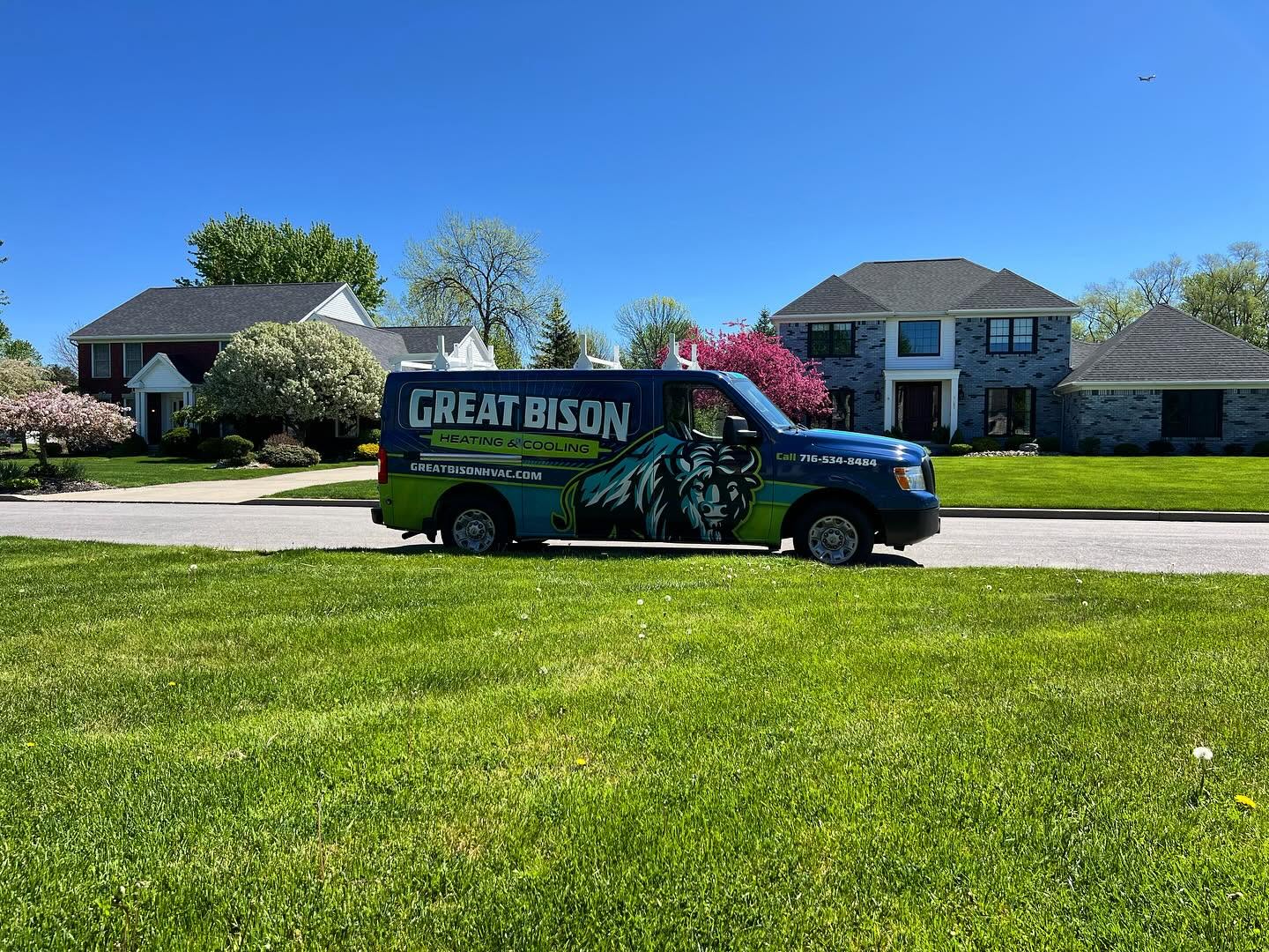 A Great Bison Heating & Cooling van is parked on a residential street with houses and trees in the background under a clear blue sky.