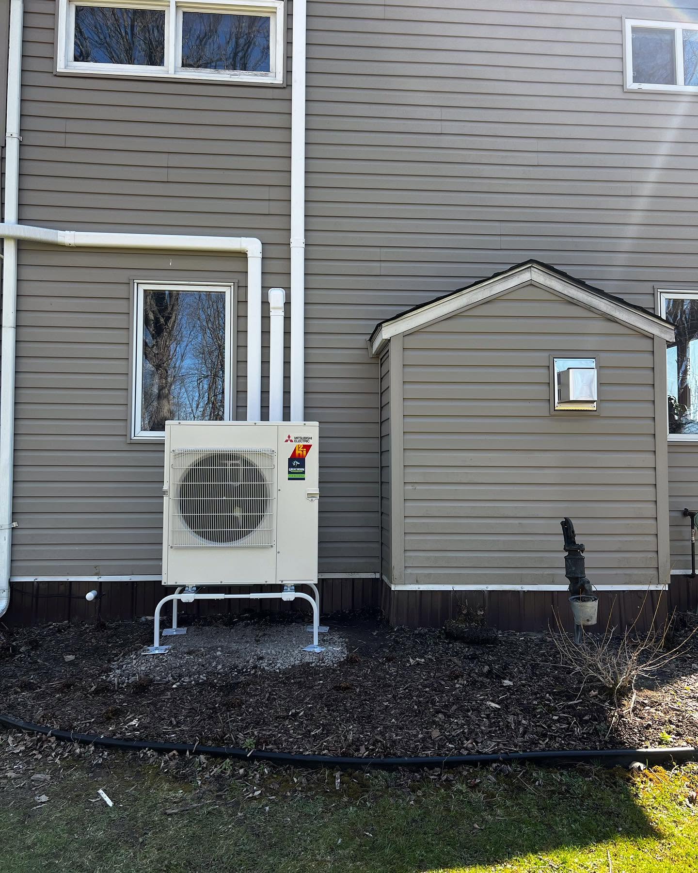 A heat pump unit is installed on a stand next to a gray house with two windows and a small utility shed. Bare soil and some pipes are visible in the foreground.