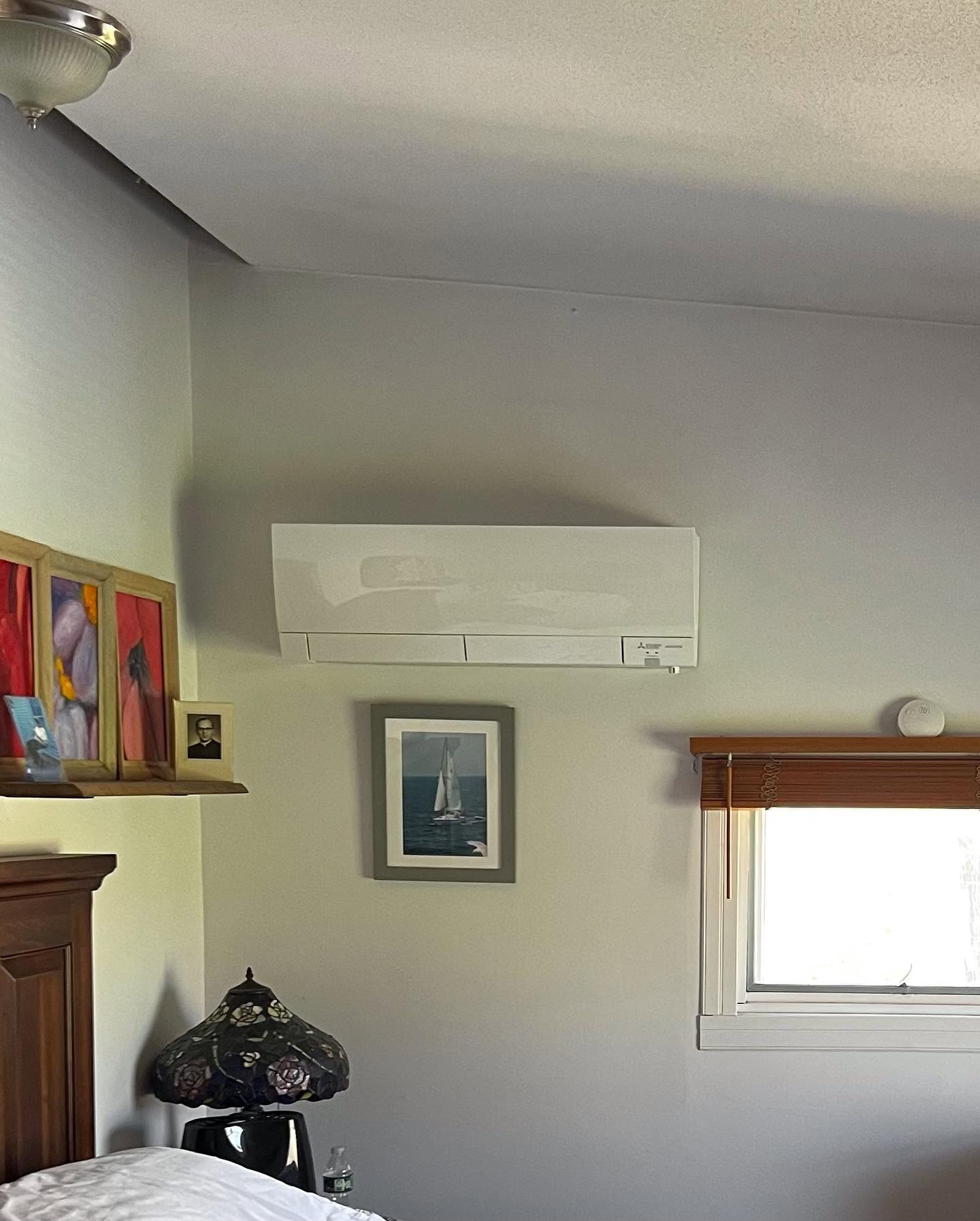 A white wall-mounted air conditioning unit is installed above a framed sailboat picture in a bedroom with a window, blinds, and decorative items.