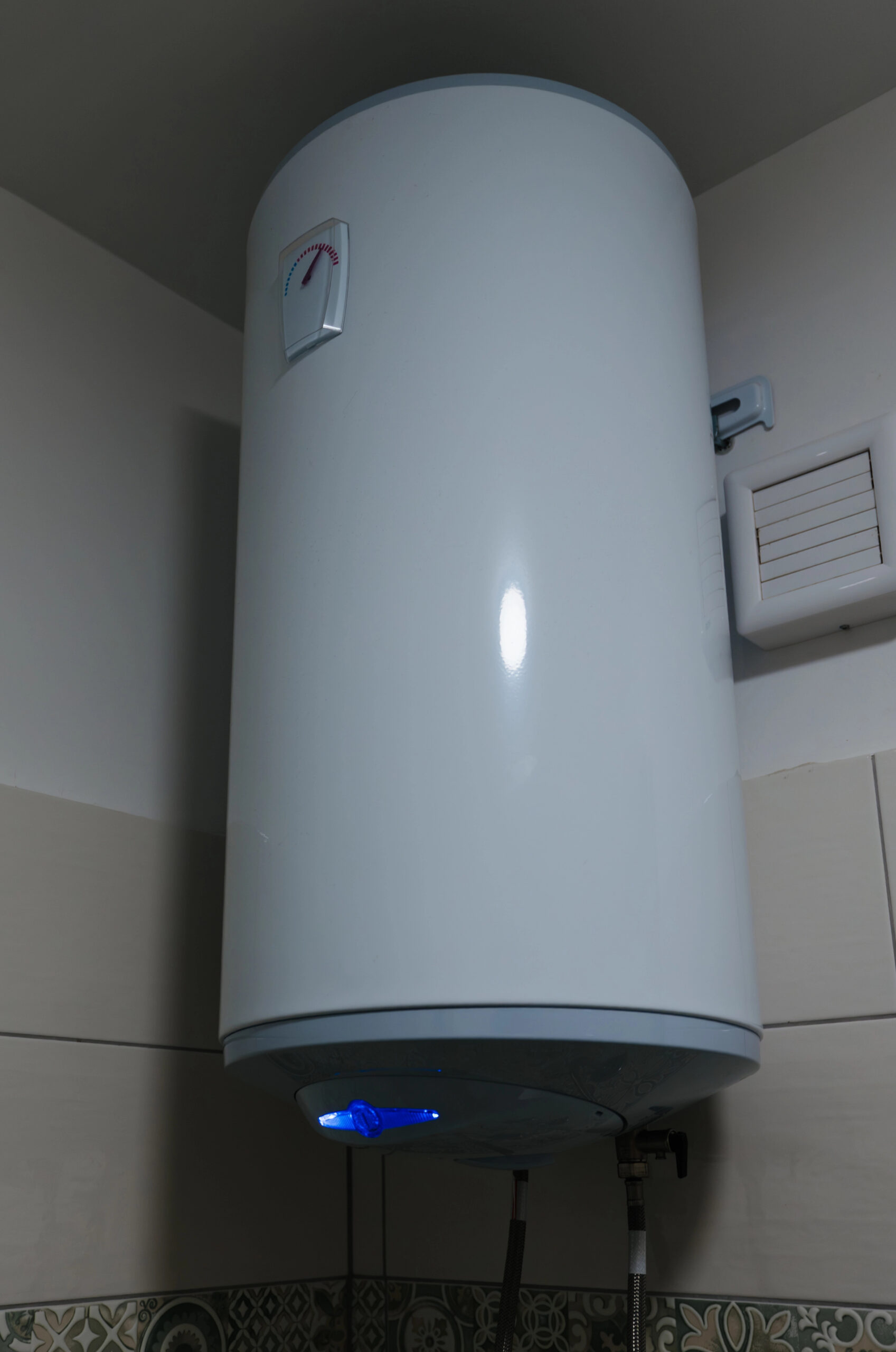 A white electric water heater is mounted on a wall in a corner, with indicator lights on at the bottom and pipes connected below.