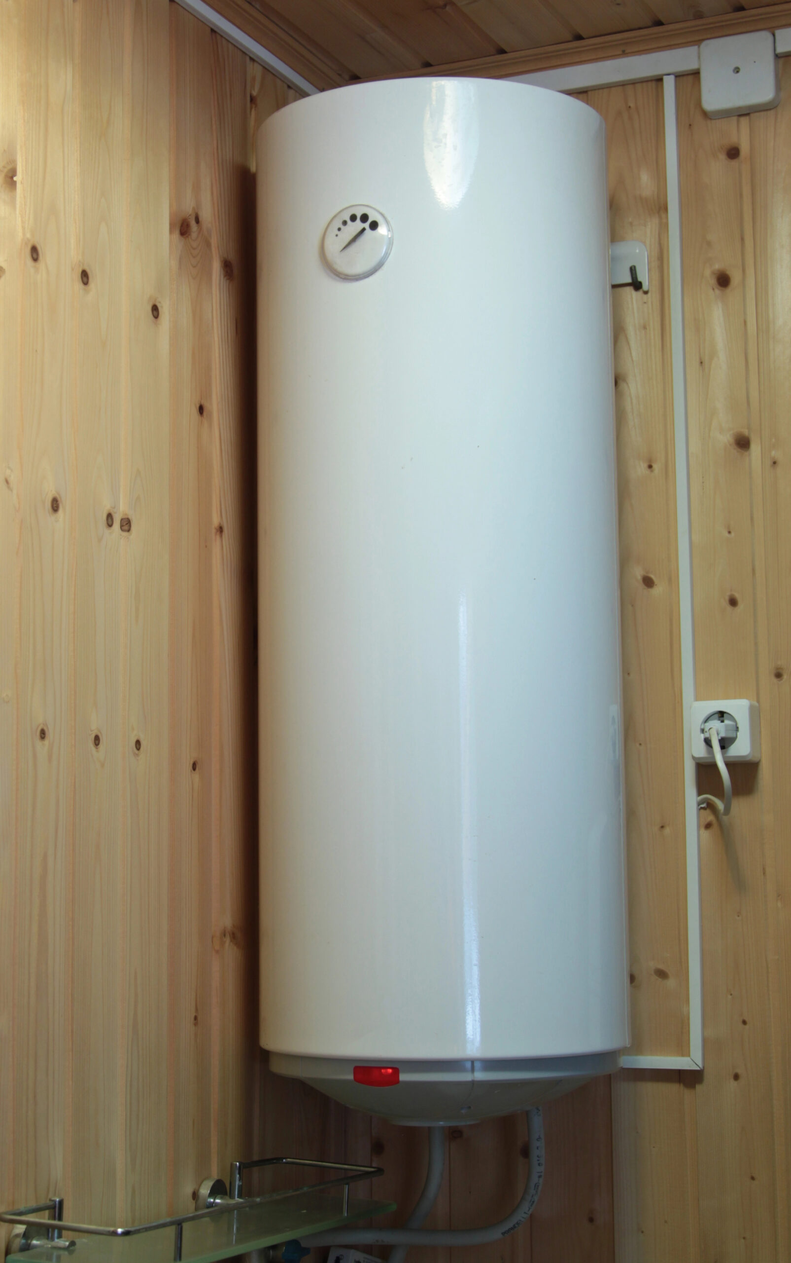 A white, cylindrical water heater mounted vertically on a wooden wall, with visible power cord plugged into an electrical outlet nearby.