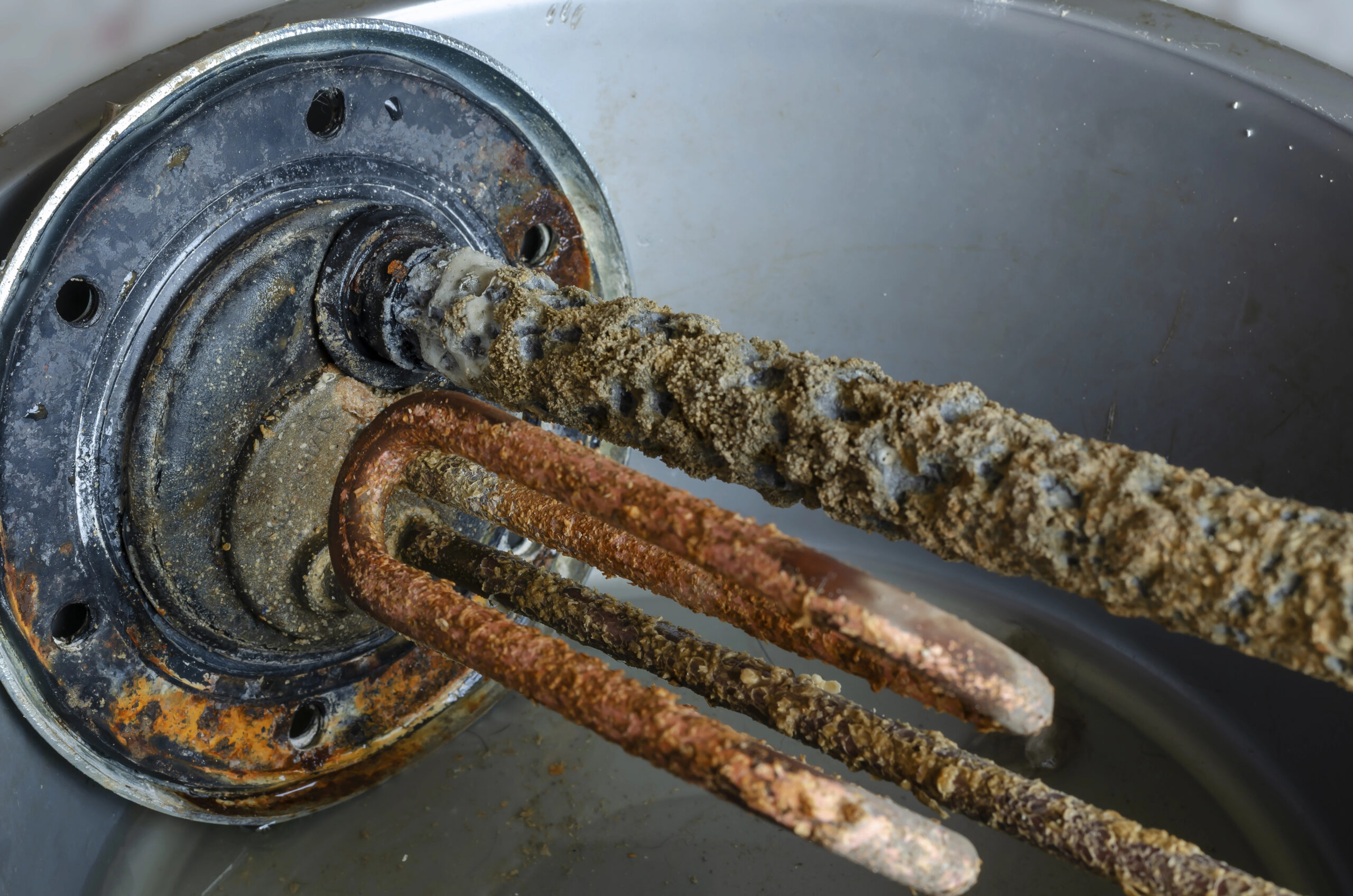 Close-up of a rusty and heavily scaled heating element inside a metal tank, showing significant mineral buildup and corrosion.