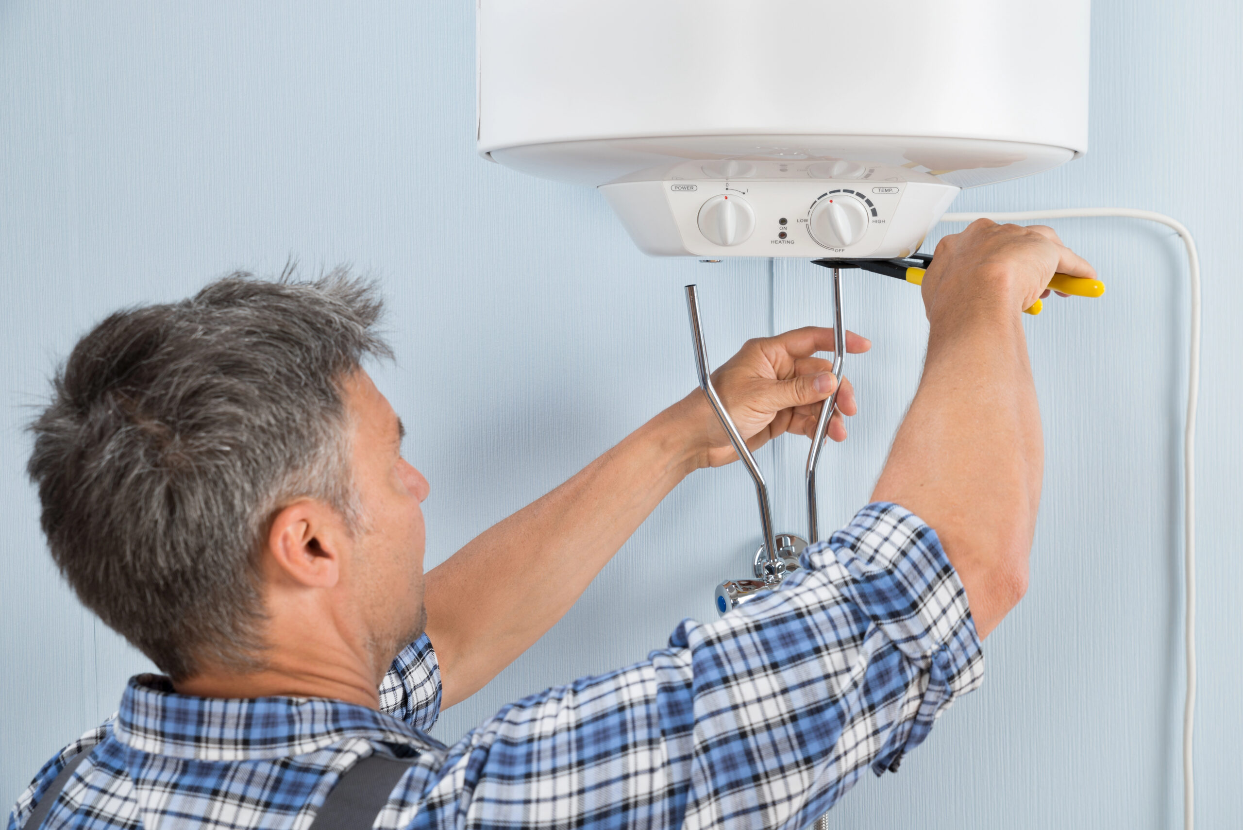 A man in a plaid shirt uses tools to repair or install a wall-mounted water heater.